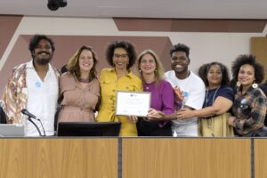 Foto mostra Dalcira recebendo a homenagem na ALMG. Ela está de pé, rodeada por deputadas e outras pessoas presentes ao evento. 